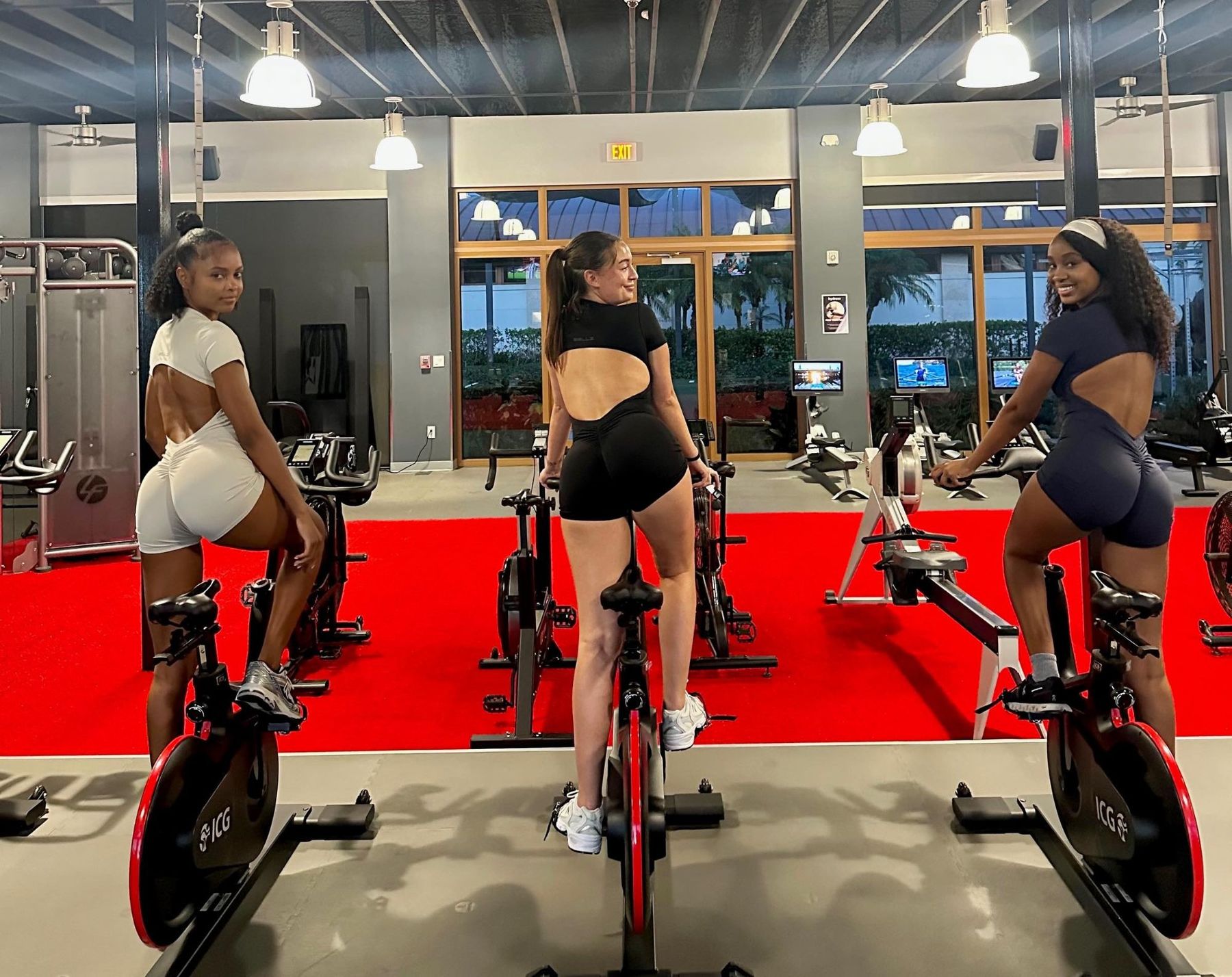 Three women on spin bikes showing the open back of Bellz bodysuits in cream, black and navy